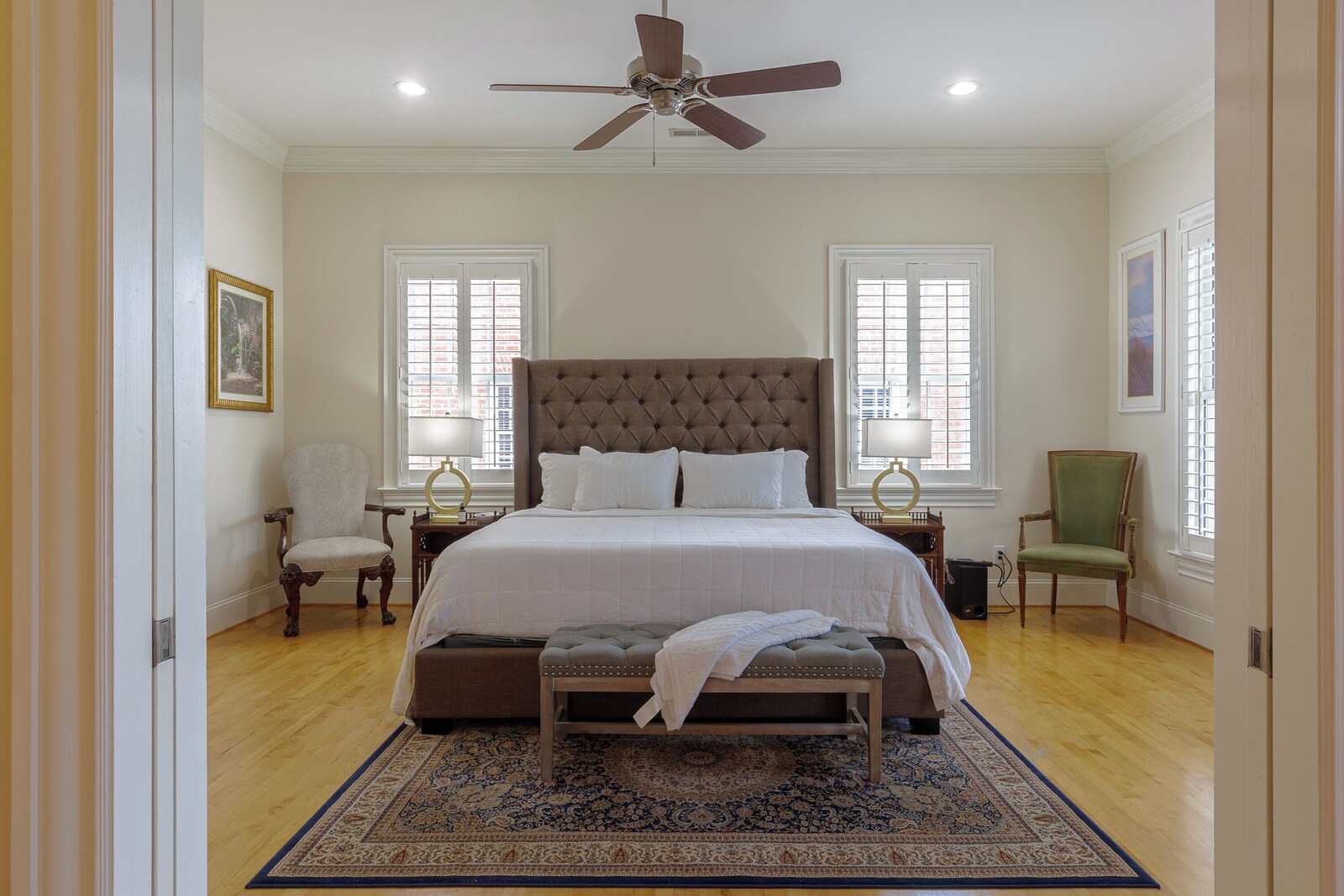 Primary bedroom seen through open double doors: king bed with a tufted gray headboard and white bedding, a tufted bench at the foot, a navy patterned rug, matching gold lamps and plantation shutters on two windows